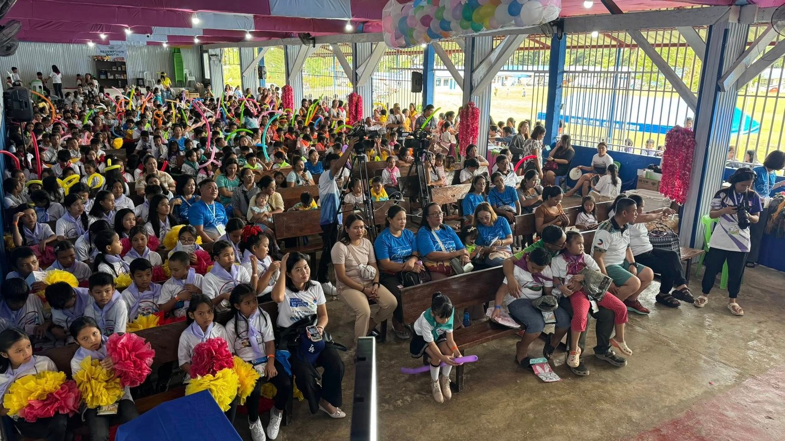 Children sitting in the auditorium, some waving the balloons